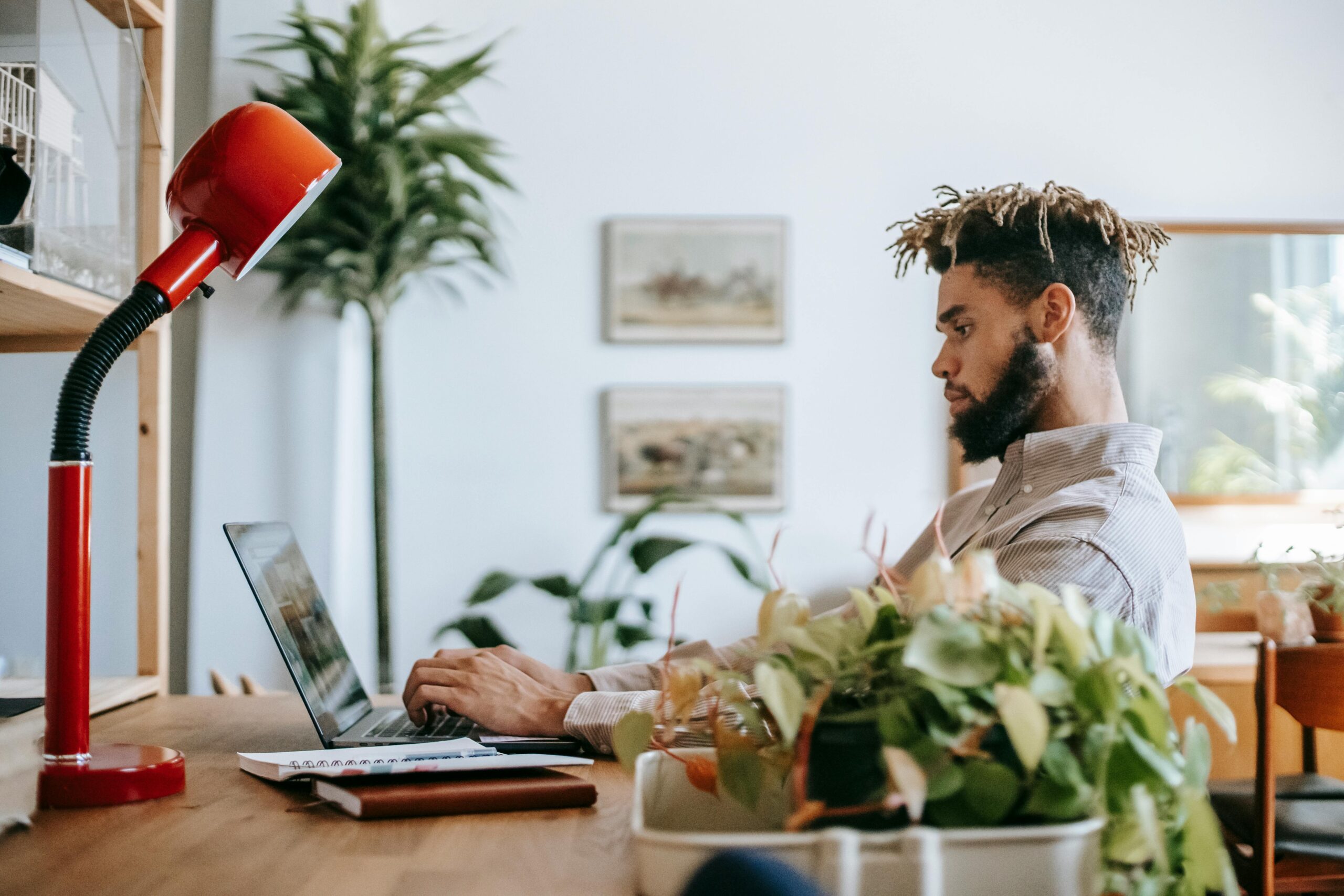 Side view of concentrated black guy in casual outfit sitting at wooden table while working remotely on computer with notepad and lamp in workspace